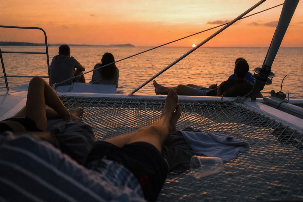 Passengers relaxing on the netting of a catamaran as the sky turns orange, no text, no logos, no people.