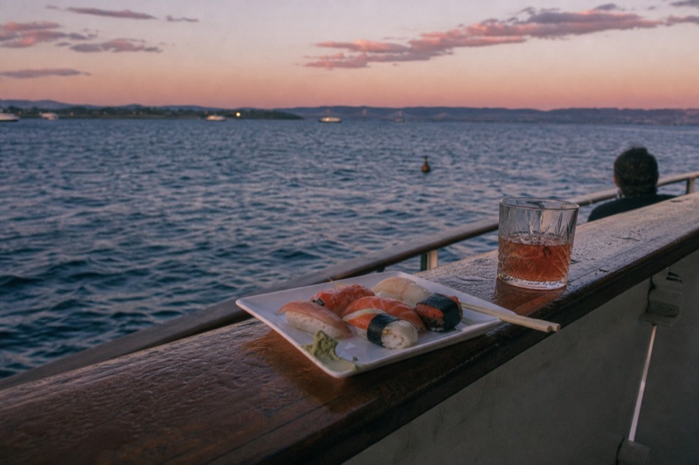 A small plate of sushi and a cocktail on a boat rail with the sea behind, no text, no logos, no people.