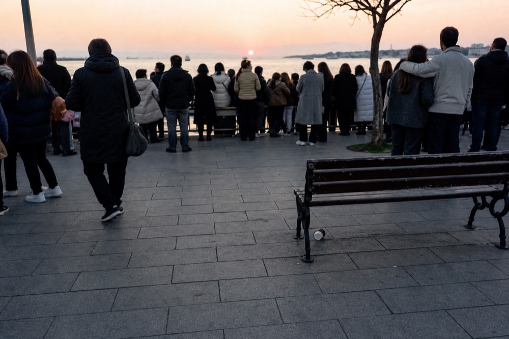 A crowded waterfront plaza at dusk with street performers and the sun low over the water, no text, no logos, no people.