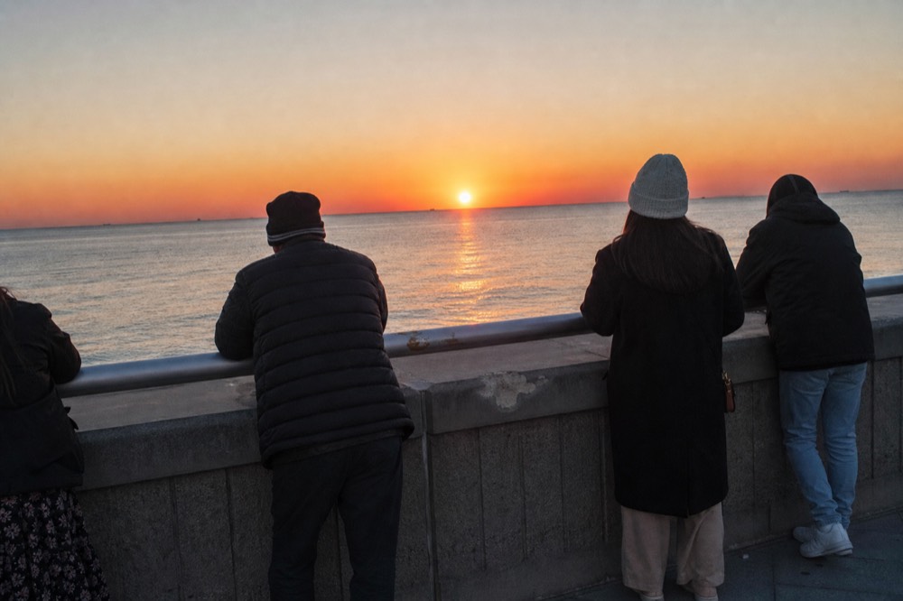 Visitors leaning on a seawall railing watching an orange sunset over calm Gulf water, no text, no logos, no people