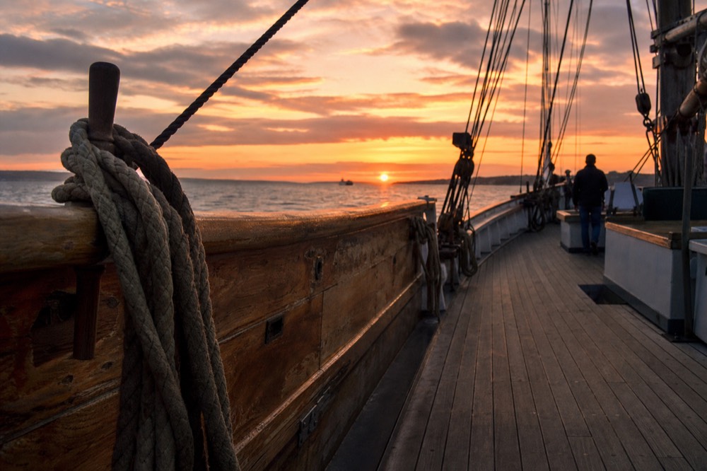 A wooden schooner deck with rigging silhouetted against an orange sunset sky, no text, no logos, no people